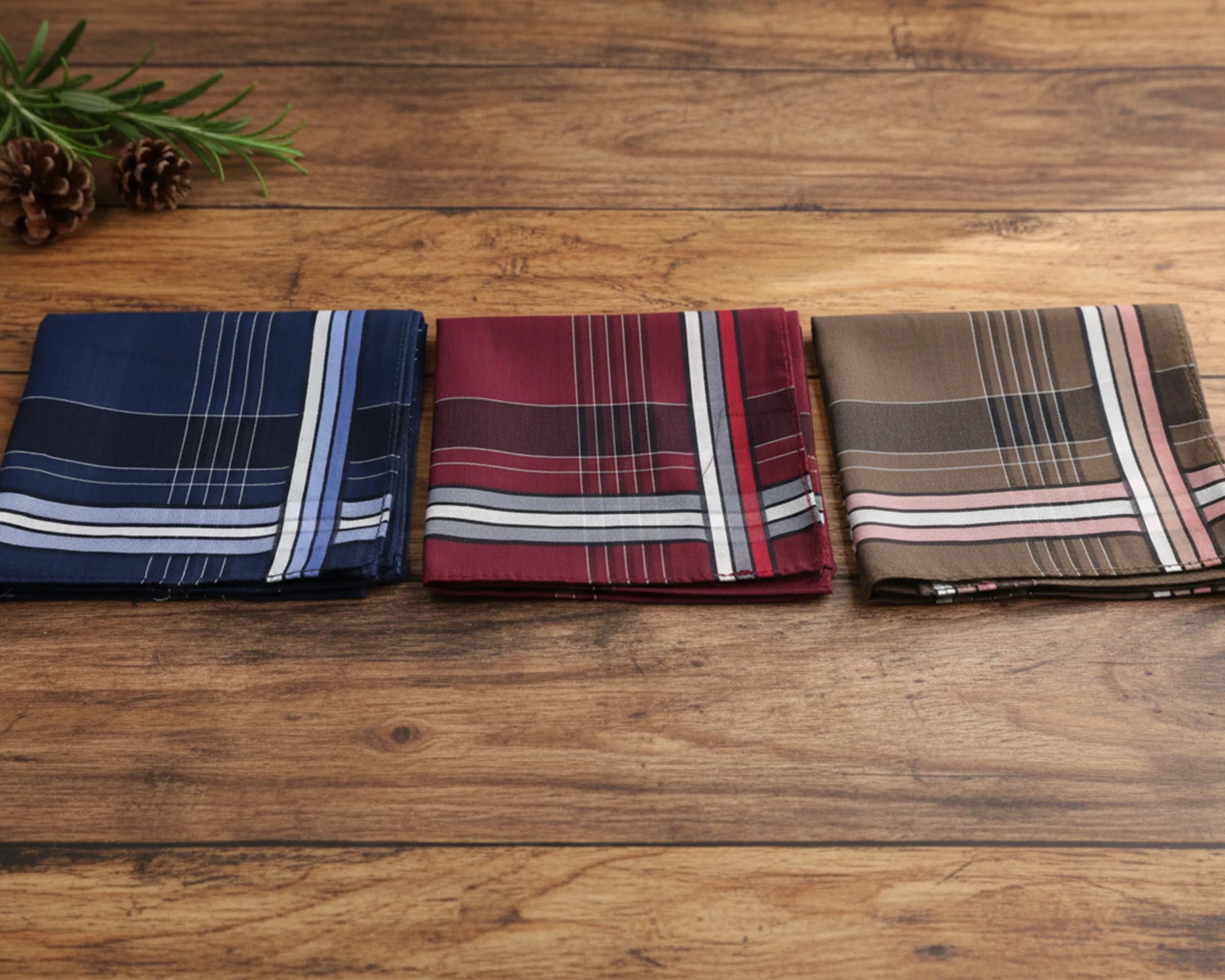 Three plaid pocket squares on a wooden surface with greenery.