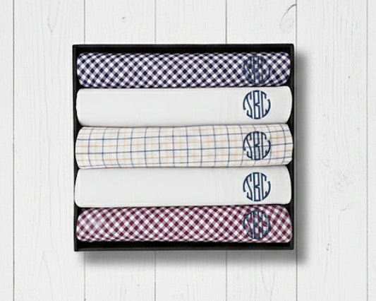 A gift box containing five folded cotton handkerchiefs in gingham and solid white with navy circular monograms.