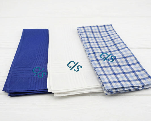 Three folded towels in blue, white, and plaid patterns with embroidered initials on a light wooden surface.