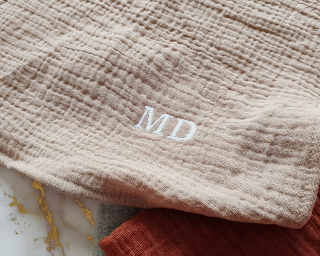 Two folded muslin cotton handkerchiefs in sage green and rust orange with navy blue JCL embroidery.