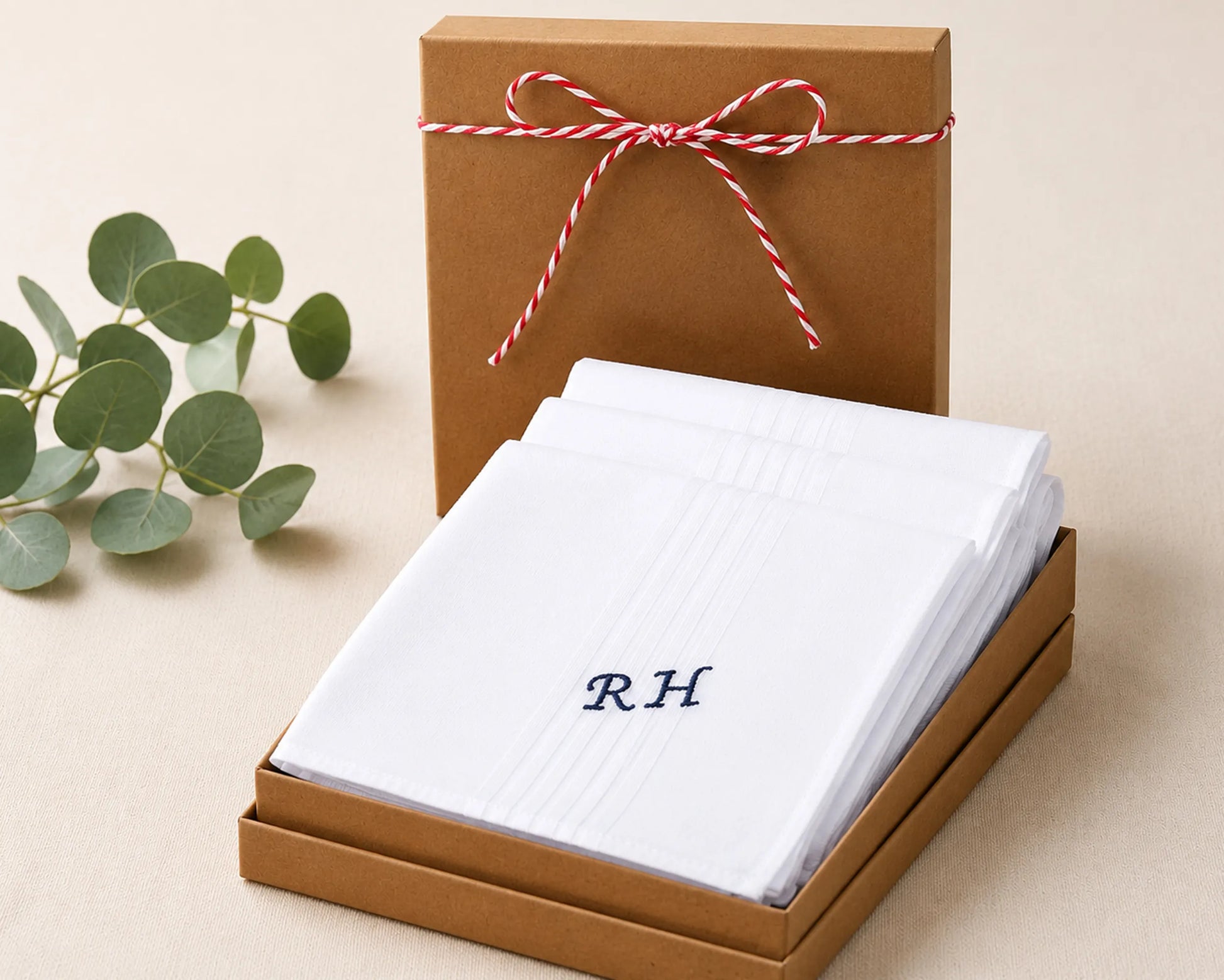 White folded fabric with initials 'RH' in a brown box with a red and white ribbon, on a beige surface with green leaves.